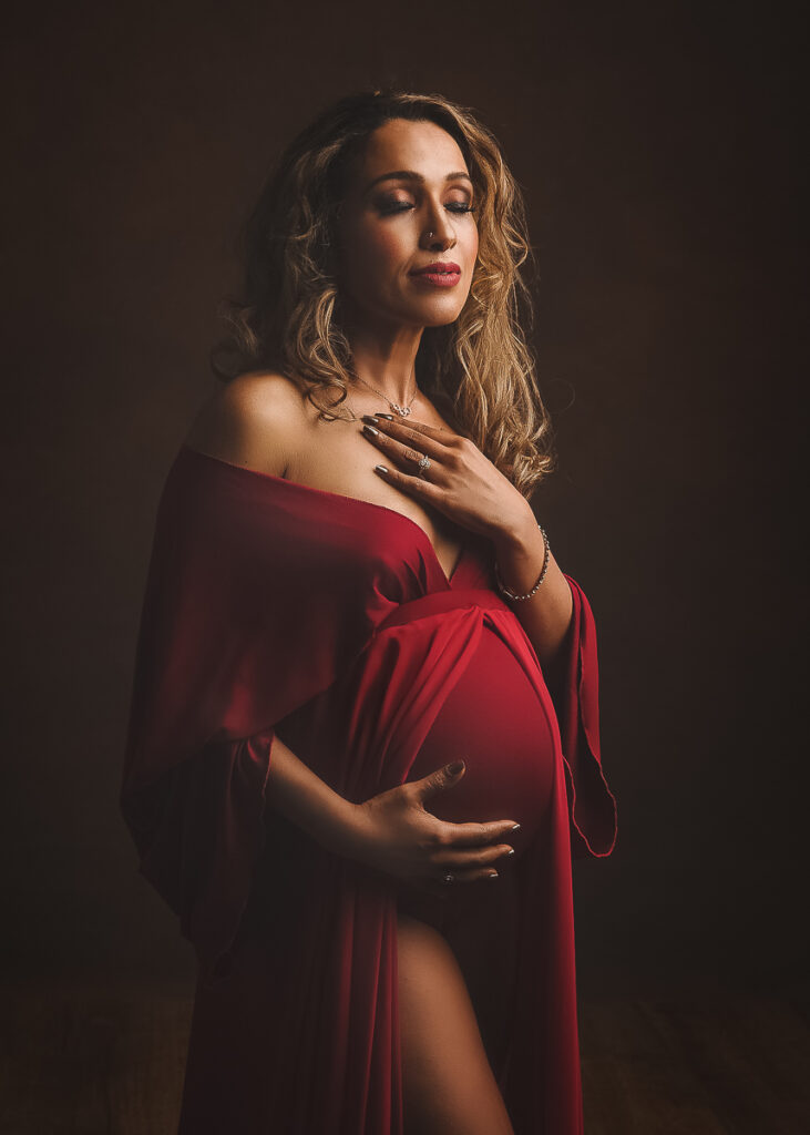 Maternity portrait of a beautiful pregnant lady wearing a loose red gown. Intimately lit and striking, showcasing her gorgeous bump. Taken at my maternity photography studio in Cheshire.